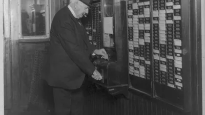 Thomas Edison, seen from behind, punching a time clock beside a wall rack of time cards in 1921